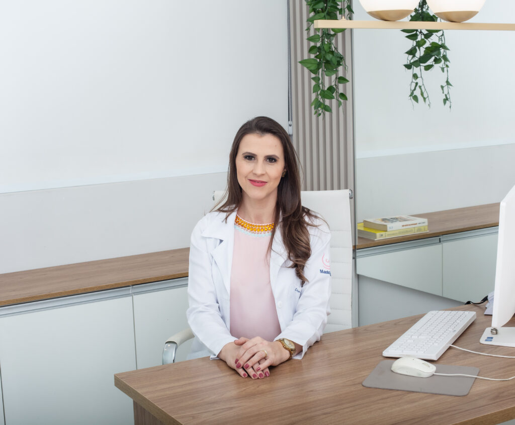 Dra. Nayara Carvalho sorri sentada à mesa do consultório, pronta para explicar o resultado da biópsia à paciente