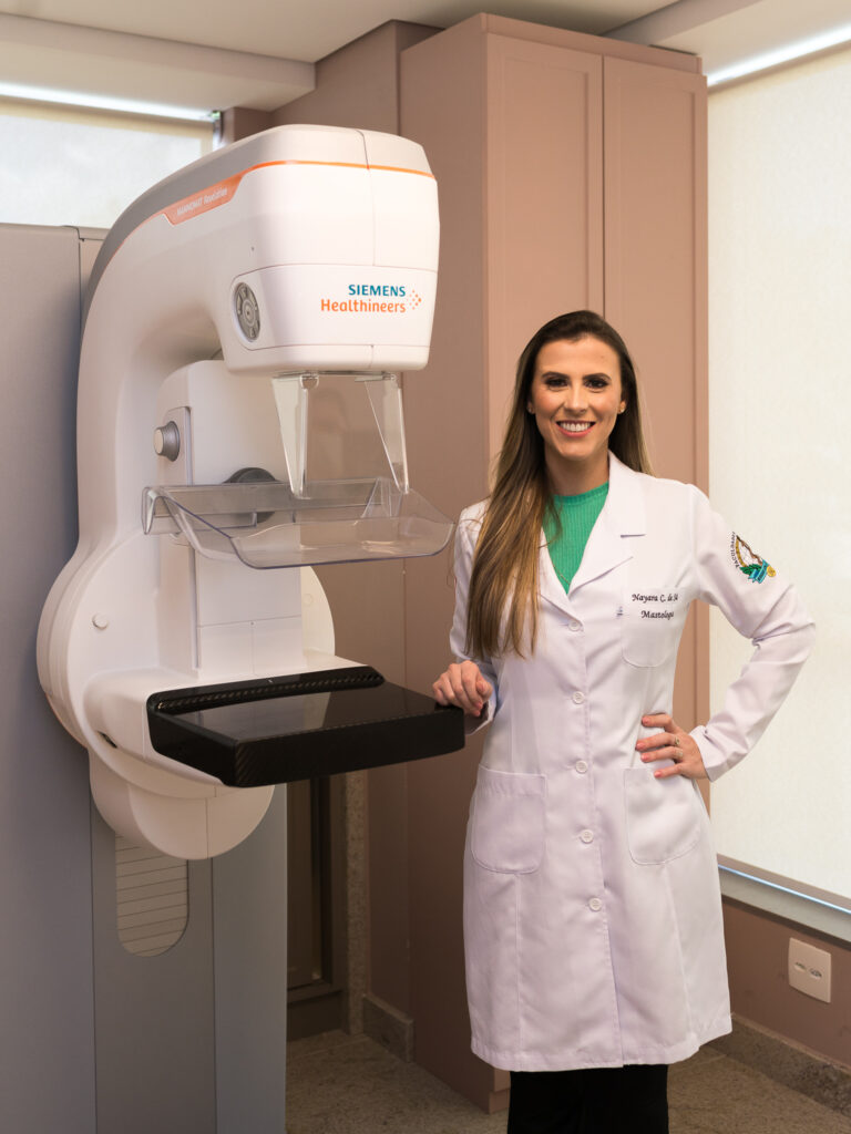 Dra. Nayara Carvalho sorri ao lado do mamógrafo Siemens, aparelho em que o clipe marcador é visualizado no acompanhamento